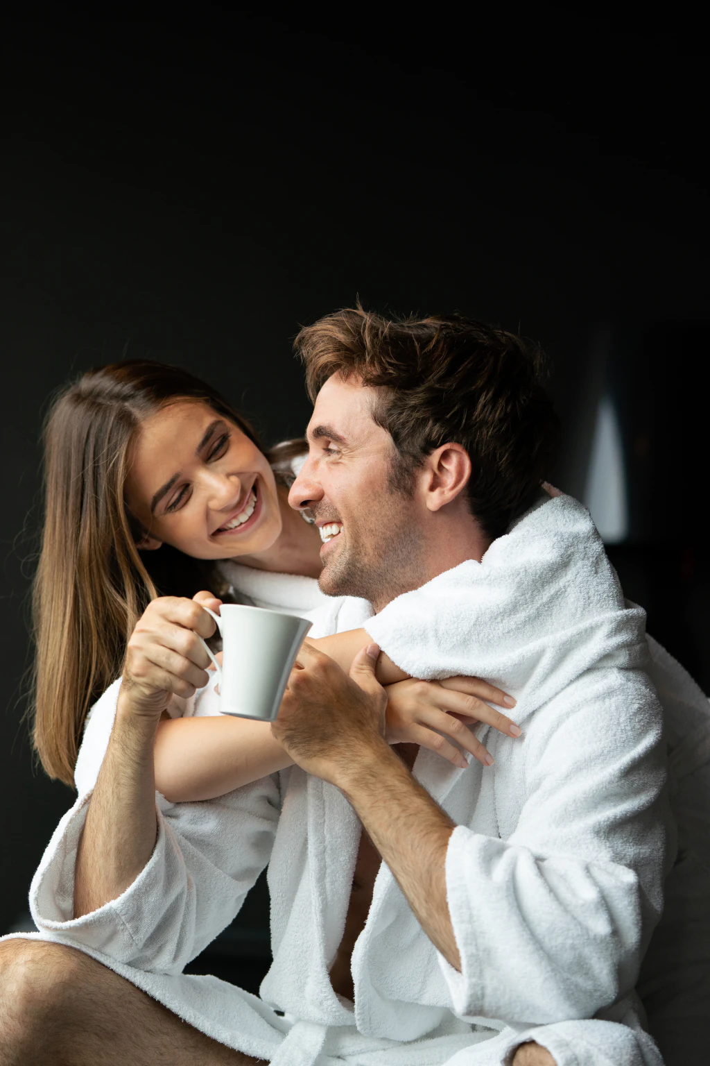Ein Paar in weißen Spa-Bademänteln umarmt sich lachend bei einer Tasse Kaffee.